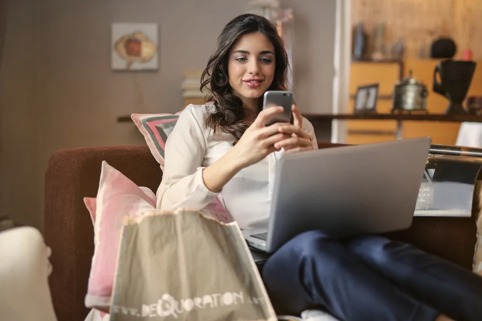 Mulher sentada no sofá com um saco de compras, a ver o telemóvel com o portátil ao colo.