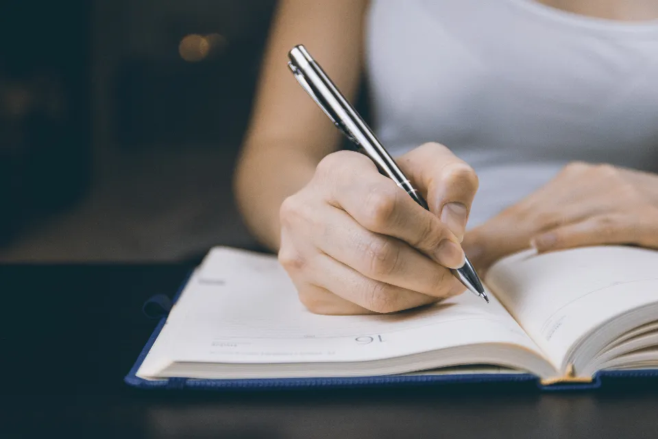 Mão a escrever num caderno aberto, como quem prepara uma reclamação.