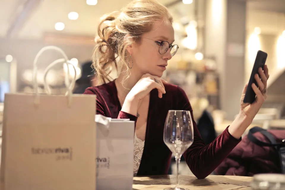Mulher com sacos de compras numa mesa, a olhar desconfiada para o telemóvel.